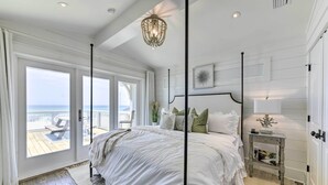 A bedroom with a large bed, a nightstand, a chandelier, and a view of the ocean through large windows.