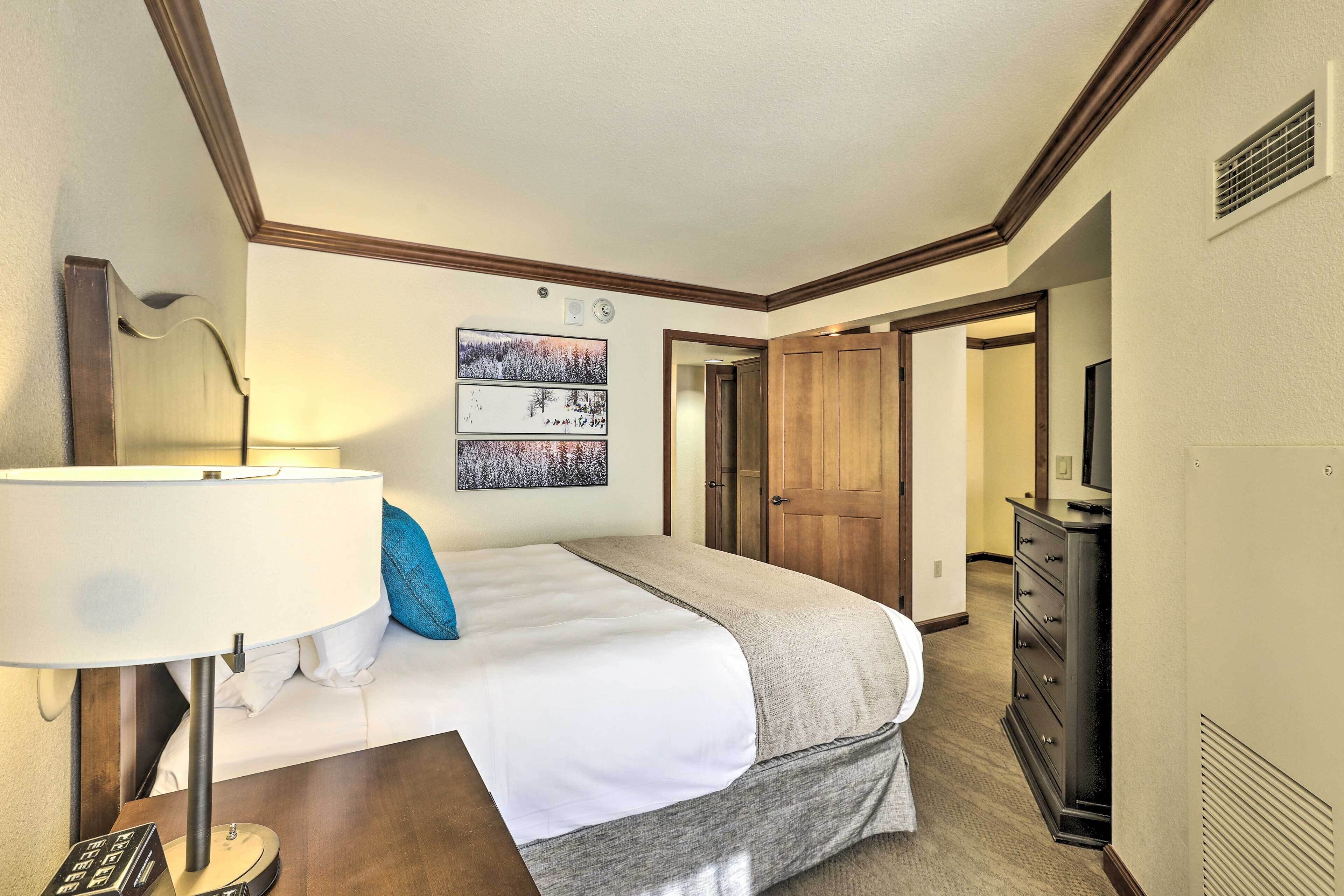 Une chambre d’hôtel comprenant un lit, un bureau, une lampe, une commode et une porte donnant sur une autre pièce.