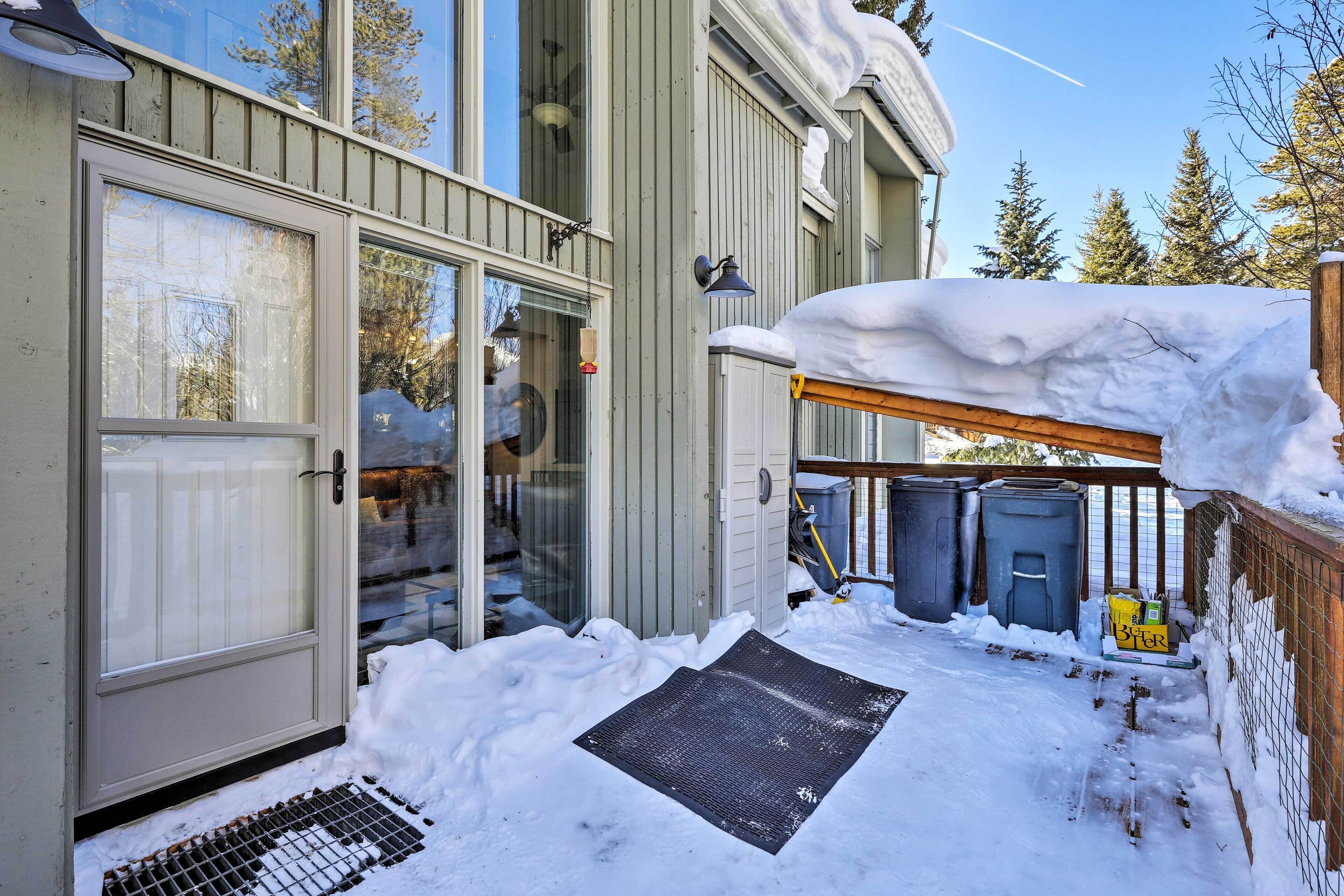 Une porte d’entrée recouverte de neige, dotée d’un panneau en verre, d’une plaque de protection en métal et d’une terrasse en bois avec une rambarde en métal.