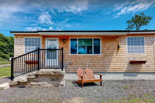 Una casa de madera de dos pisos, con un porche delantero, una escalera de piedra y una silla Adirondack de madera.