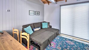 Ein Zimmer mit Holzbalkendecke, einem grauen Sofa mit türkisfarbenen Kissen, einem Holztisch und einem großen Fenster mit weißem Rollo.