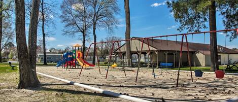 Un parque infantil con columpios y tobogán, rodeado de árboles y un edificio al fondo.