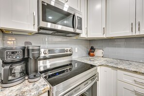 A kitchen with white cabinets, a stainless steel microwave, a Keurig coffee maker, an oven, and a granite countertop. - Elegant FL Condo w/ Pool: Walk to Madeira Beach! (Madeira Beach)