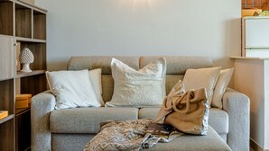 Ein Wohnzimmer mit grauem Sofa, zwei weißen Kissen, einer gemusterten Decke und einer beigen Tasche. Eine Regalwand mit Dekoration ist links davon.