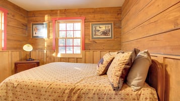 Una habitación con paneles de madera, una cama, una mesita de noche, una ventana con detalles rojos y dos cuadros en la pared.