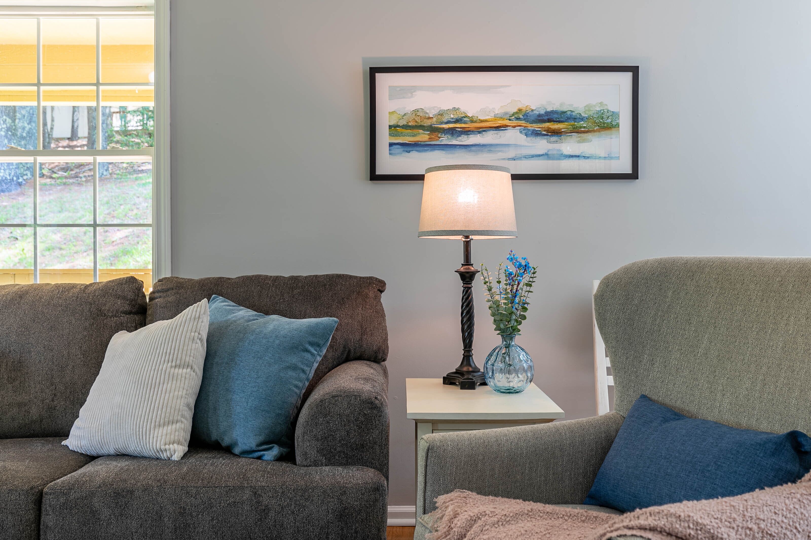 A living room with a grey sofa, a blue armchair, a small table with a lamp, and a framed landscape painting on the wall.