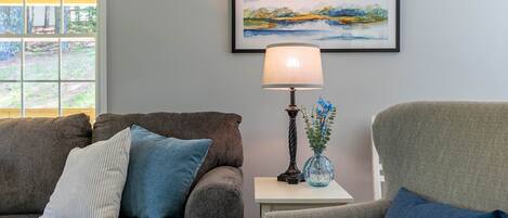 A living room with a grey sofa, a blue armchair, a small table with a lamp, and a framed landscape painting on the wall.