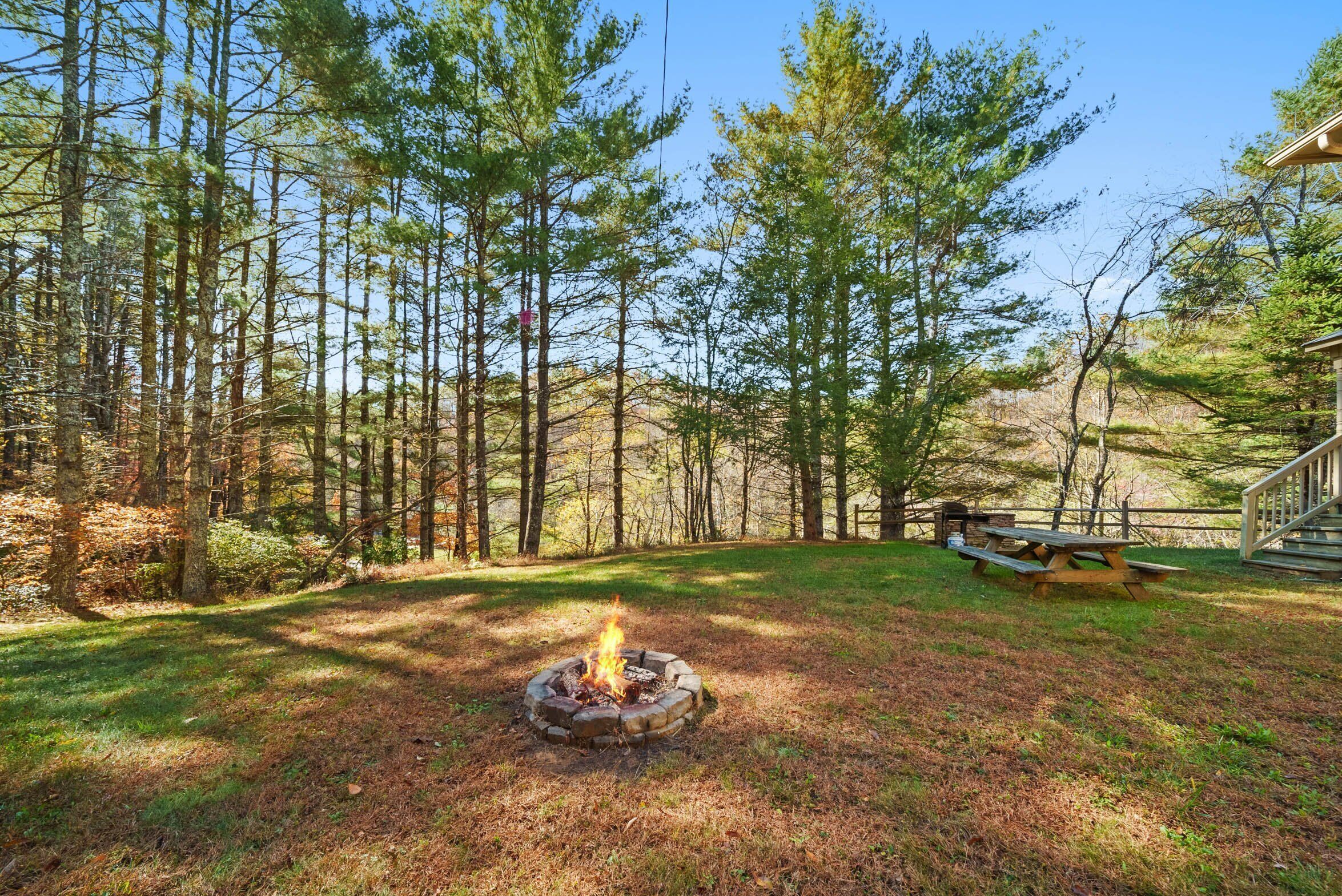 A campfire surrounded by tall pine trees in a grassy area with a picnic table and a wooden deck.