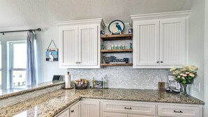 A kitchen with white cabinetry, granite countertops, and a backsplash. There are shelves with decorative items and a window with curtains. - Surfside Beach Home w/ Views, Steps to Shore! (Freeport)