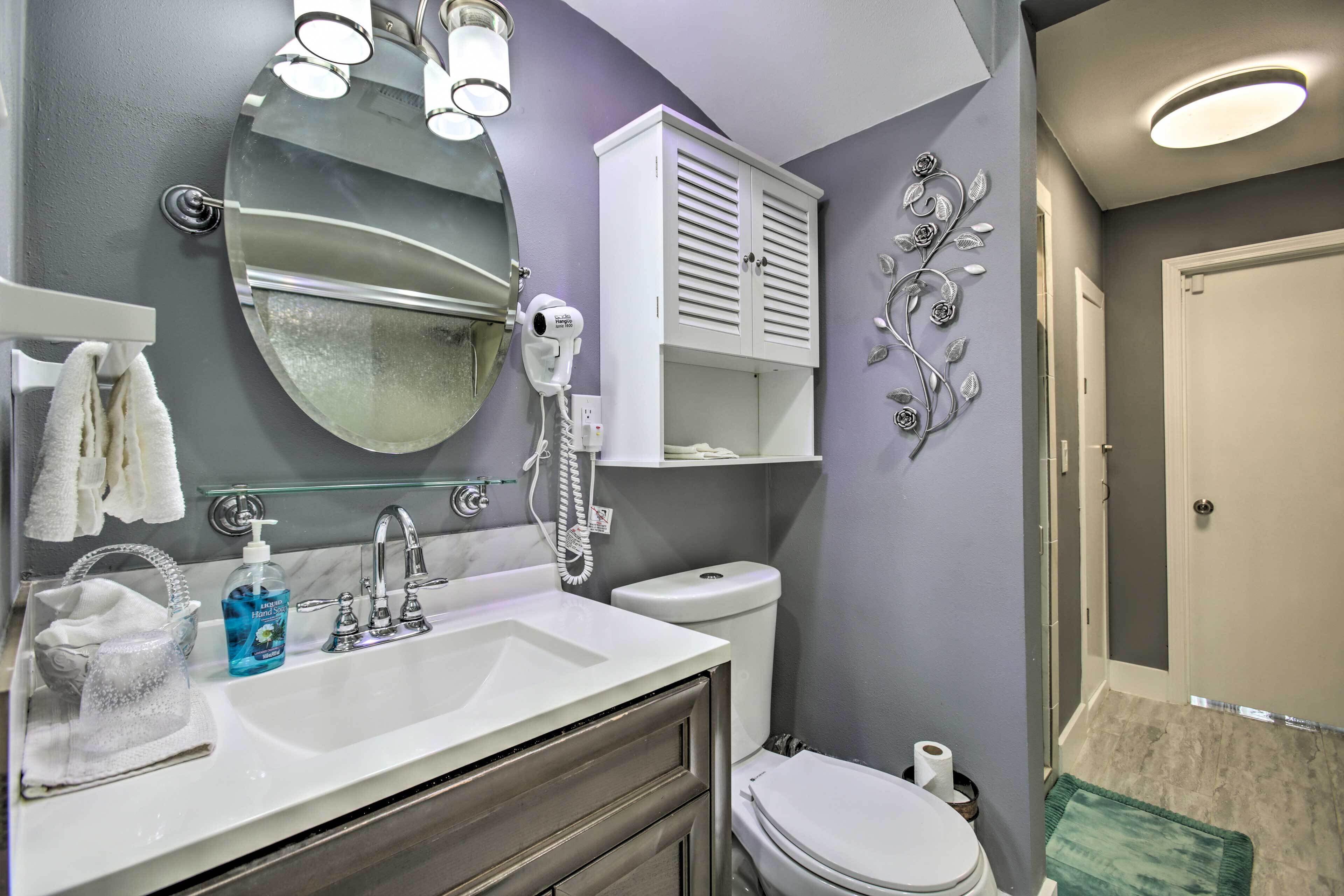 Une salle de bain avec un lavabo blanc, un miroir et des toilettes. On y trouve un sèche-cheveux et un flacon de savon liquide sur le lavabo.