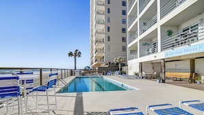 A pool area with blue and white lounge chairs, a small pool, and a building with a sign reading "Carolina Shores". - Cozy Beachfront Condo w/ Wifi, Views & Pool Access (Murrells Inlet)