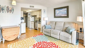 A modern living room with a sofa, a small table, and a kitchen in the background. - Cozy Beachfront Condo w/ Wifi, Views & Pool Access (Murrells Inlet)