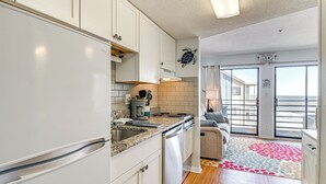 A modern kitchen with white cabinets, a granite countertop, and a dishwasher. There is a view of the ocean through large sliding glass doors. - Cozy Beachfront Condo w/ Wifi, Views & Pool Access (Murrells Inlet)