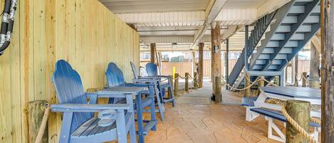 A covered outdoor seating area with wooden walls and stone flooring, featuring blue chairs and tables, and a staircase leading to an upper level.