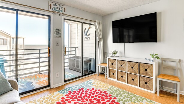 A living room with a large sliding glass door leading to a balcony, a flat-screen TV mounted on the wall, a wooden cabinet with storage boxes, and a colorful rug. - Cozy Beachfront Condo w/ Wifi, Views & Pool Access (Murrells Inlet)