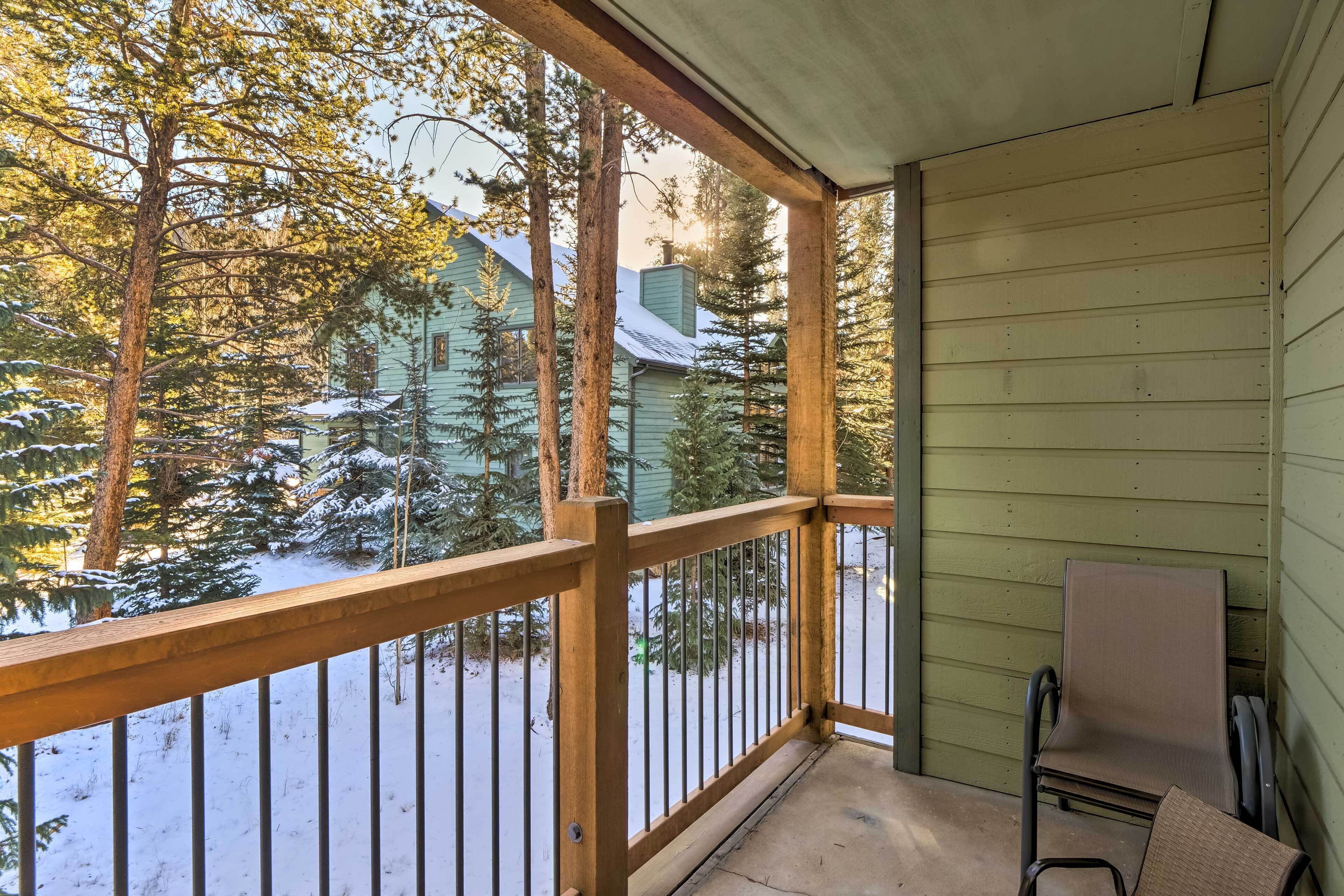 A covered porch with a wooden railing, a chair, and a view of snow-covered trees and houses.