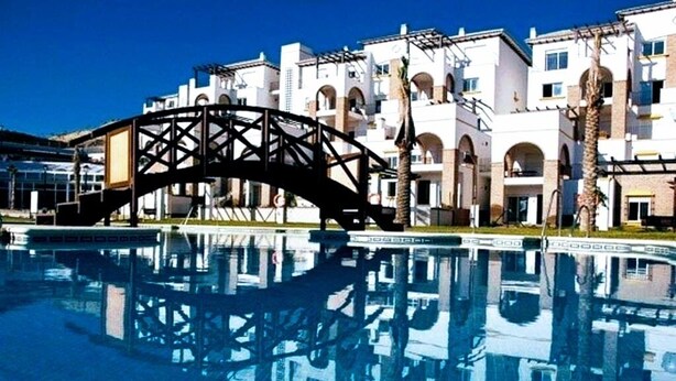 A swimming pool with a bridge structure in the foreground and a row of white buildings with arched windows in the background.