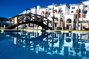 A swimming pool with a bridge structure in the foreground and a row of white buildings with arched windows in the background.