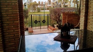 A view from a balcony with a glass floor, a brick wall, and a metal gate leading to a courtyard with a small dog statue, a grill, and a grassy area with buildings in the background.