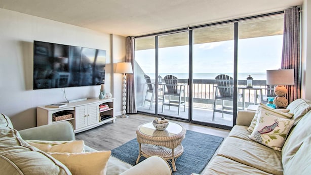 A modern living room with a large TV, comfortable seating, a small round table, and a balcony with a view of the sea. - Indoor Pool & Views: Beachfront Ocean City Condo (Ocean City)
