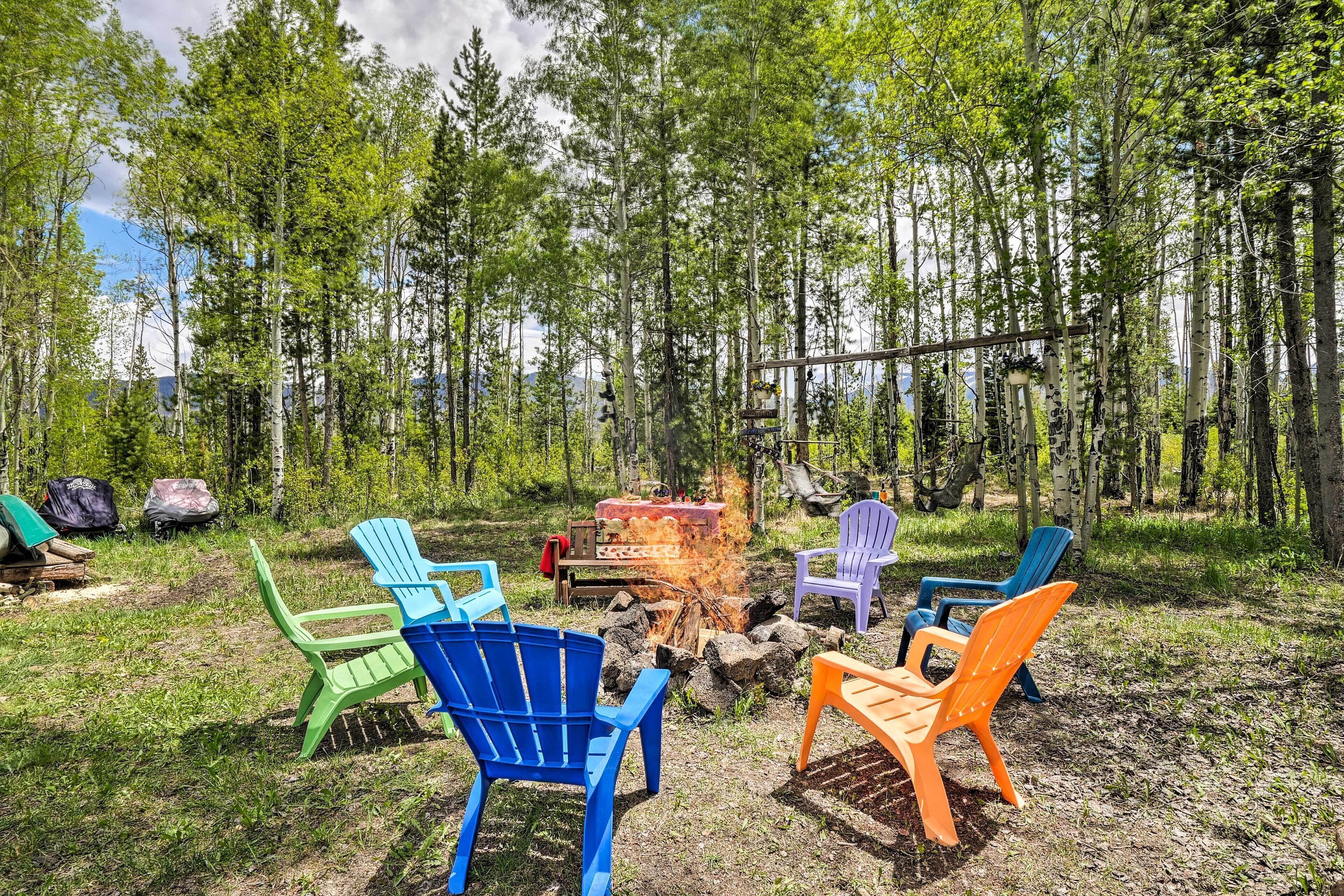 Ein Lagerfeuer mit bunten Adirondack-Stühlen, die darum herum in einer Waldlandschaft angeordnet sind.