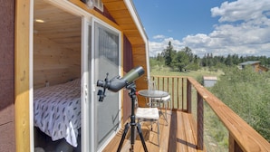 A wooden cabin with a telescope on a balcony, a bed with a leaf-patterned bedspread, and a small table and chair. - Secluded Jefferson Cabin w/ Deck + Mountain Views! (Jefferson)
