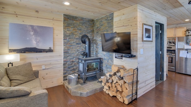 A cozy living area with a stone fireplace, a television, a sofa, and a kitchen area with modern appliances. - Secluded Jefferson Cabin w/ Deck + Mountain Views! (Jefferson)