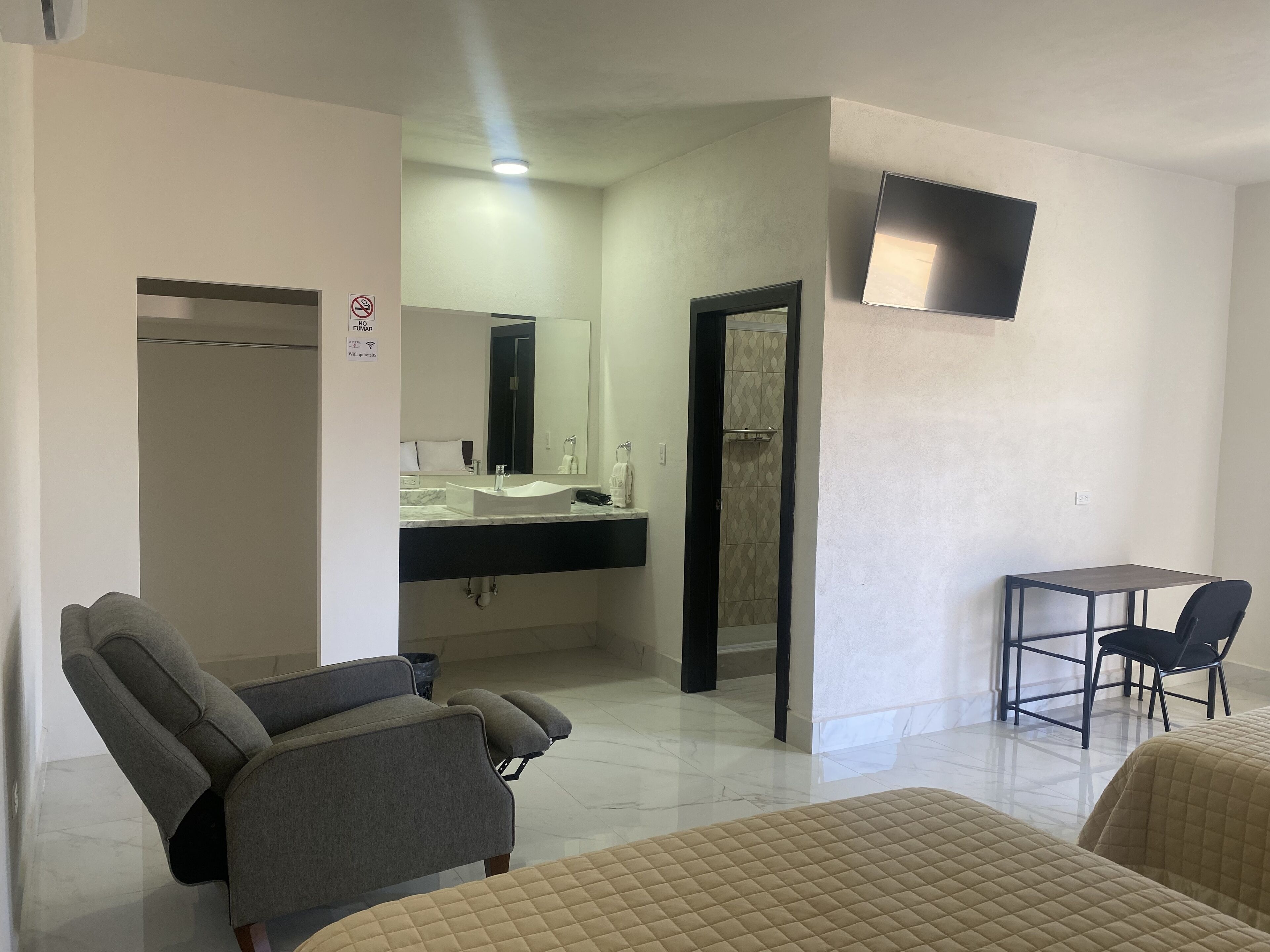 Une chambre d’hôtel comprenant un canapé, un lit, une petite table avec des chaises, une salle de bain avec un lavabo et un miroir, et un téléviseur fixé au mur.