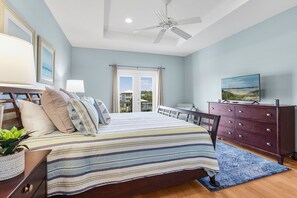 Quarto com cama, cômoda, TV, ventilador de teto e janela com cortinas.