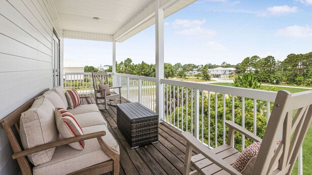 A covered porch with a wooden bench and cushions, a storage box, and a view of a green landscape with trees and a house in the distance. - Hangloose Hideaway Beach House w/ a pool. 1 block to beach. Great fall rates! (Gulf Shores)