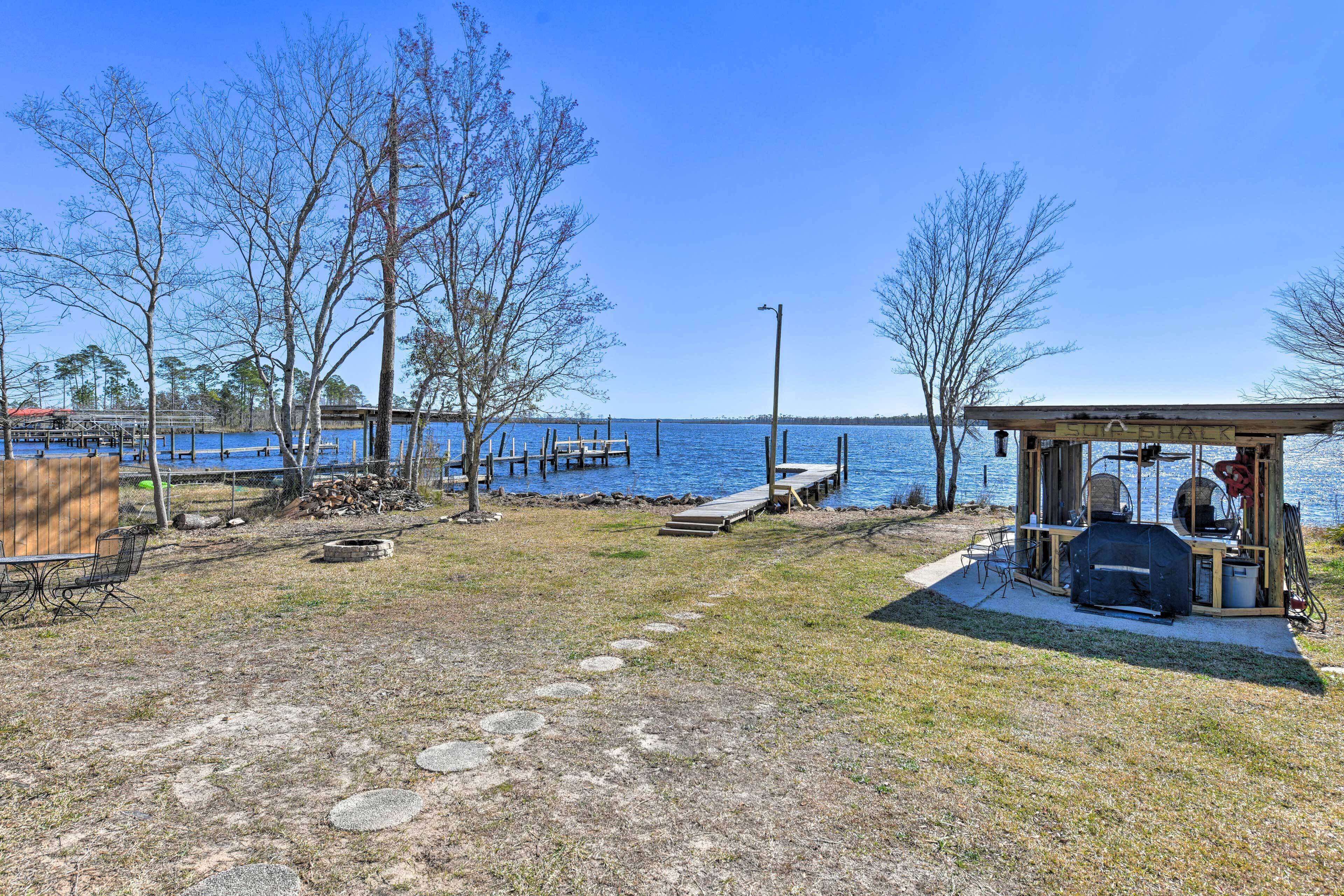 A lakeside outdoor area with a barbecue setup, a covered seating area, and a dock extending into the water.