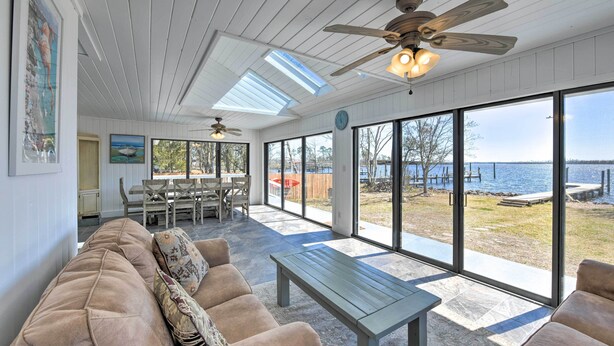 A spacious living area with a large window overlooking a body of water, a ceiling fan, and a dining area with a table and chairs. - Updated Waterfront Escape w/ Dock & Fire Pit (Pensacola)