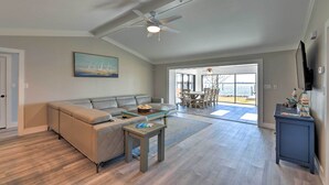 A spacious living room with a beige sectional sofa, a coffee table, and a ceiling fan. There is a large window providing a view of the outdoors. - Updated Waterfront Escape w/ Dock & Fire Pit (Pensacola)