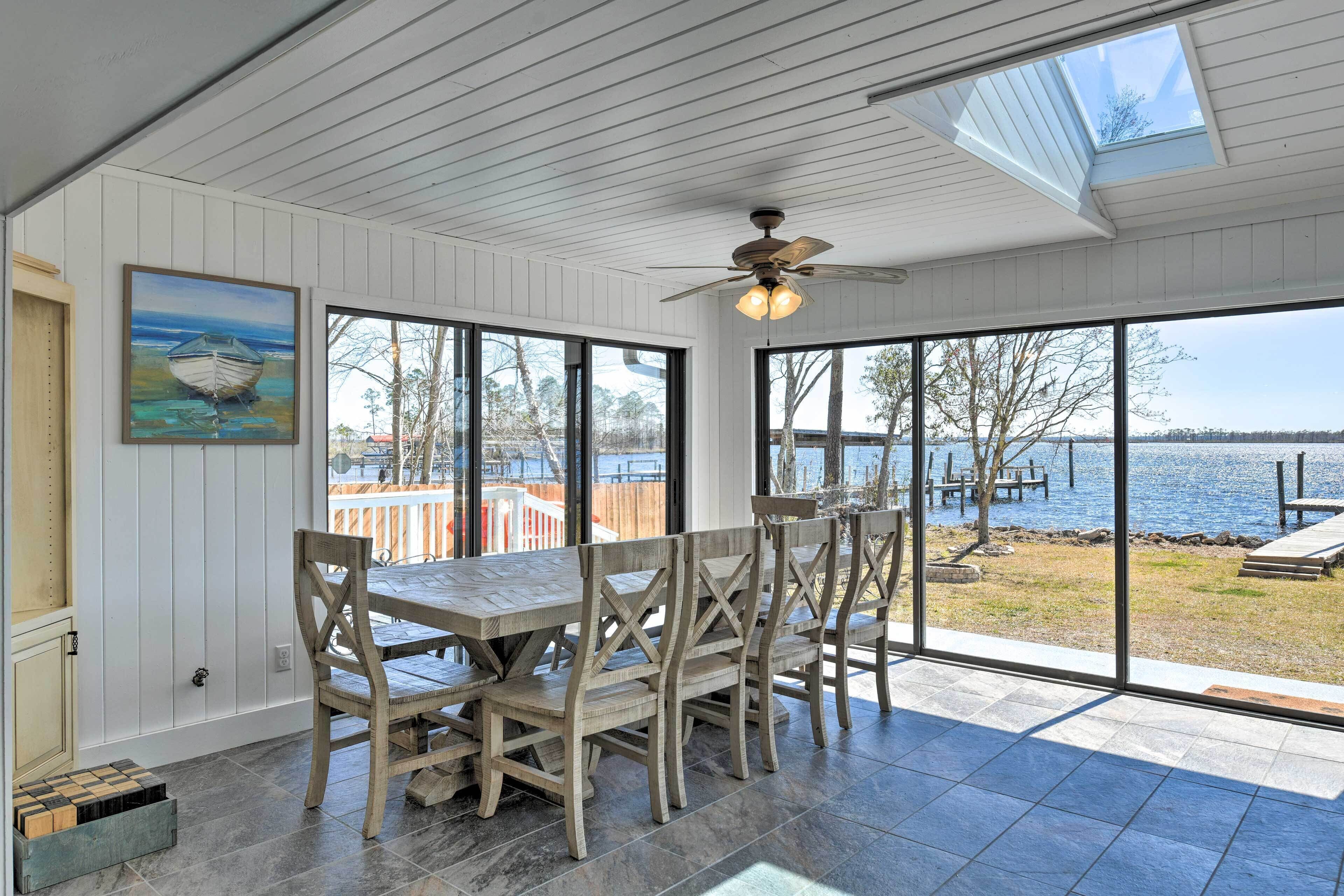 A dining area with a wooden table and chairs, large windows with a view of a lake, and a ceiling fan.