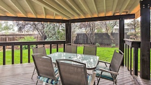 A covered outdoor patio with a glass table and chairs, surrounded by a wooden deck and railing. - Arvada Home w/ Deck & Game Room Near Olde Town! (Arvada)