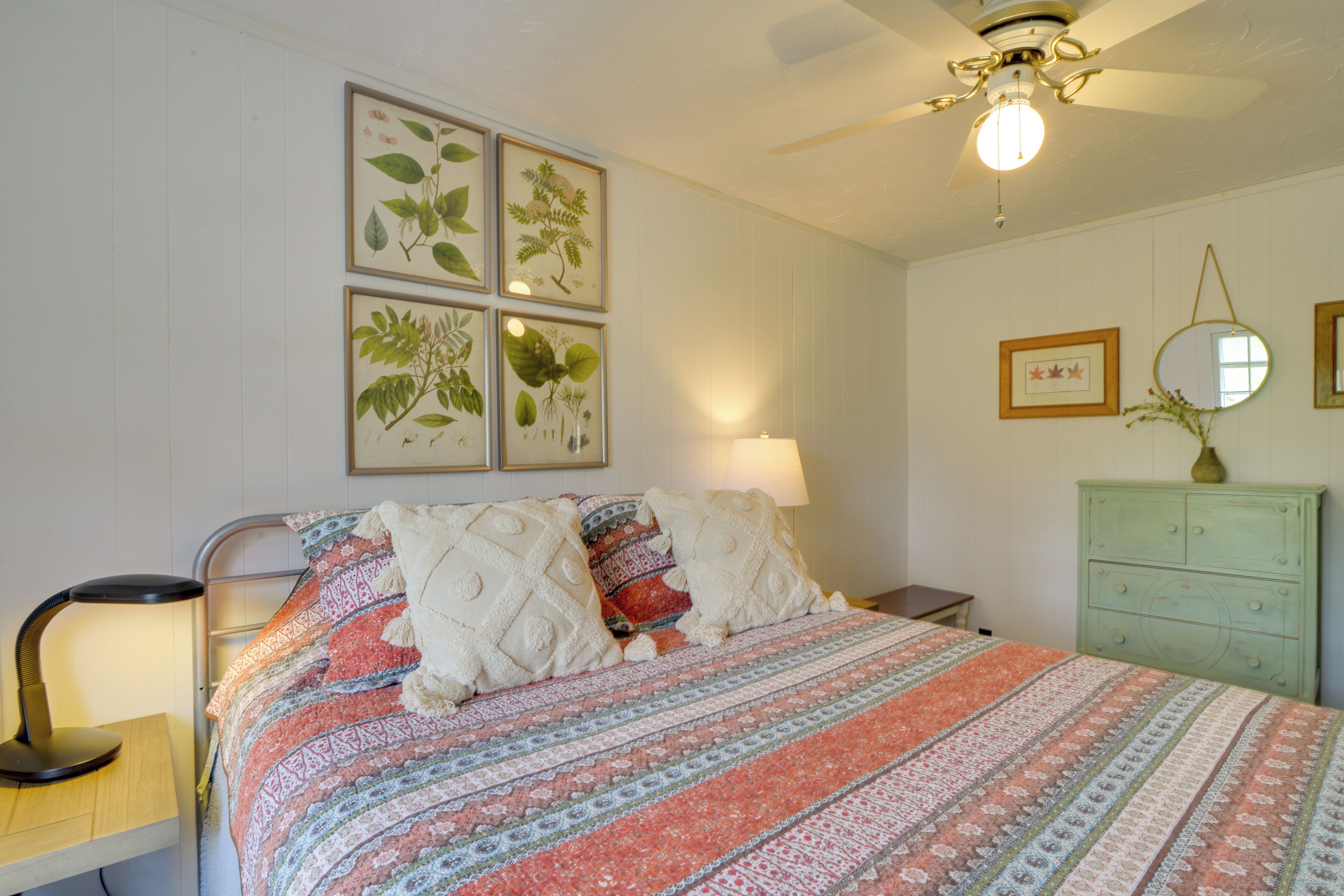 Una cama bien hecha con ropa de cama estampada, una lámpara de noche, un aparador verde e impresiones botánicas enmarcadas en la pared.