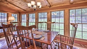 Un comedor con una mesa de madera dispuesta para seis personas, rodeada de sillas de madera, ventanas amplias y una araña.