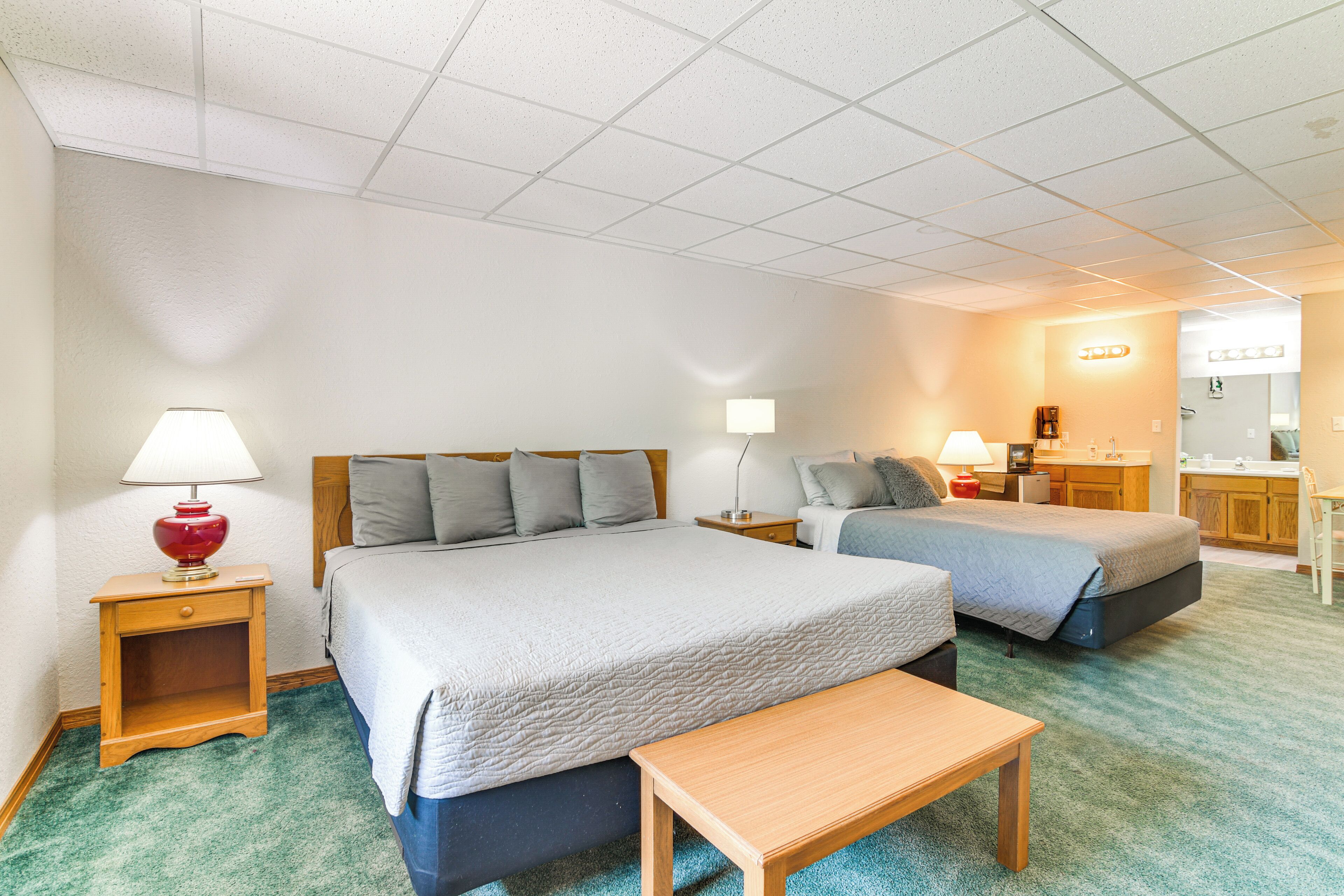 A hotel room with two beds, a wooden nightstand with a lamp, a small table, and a kitchenette area.