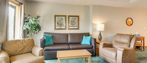 A living room with a brown leather sofa, a beige armchair, a wooden coffee table, and a teal carpet. There are two framed botanical prints on the wall, a floor lamp, and a mirror on the wall.
