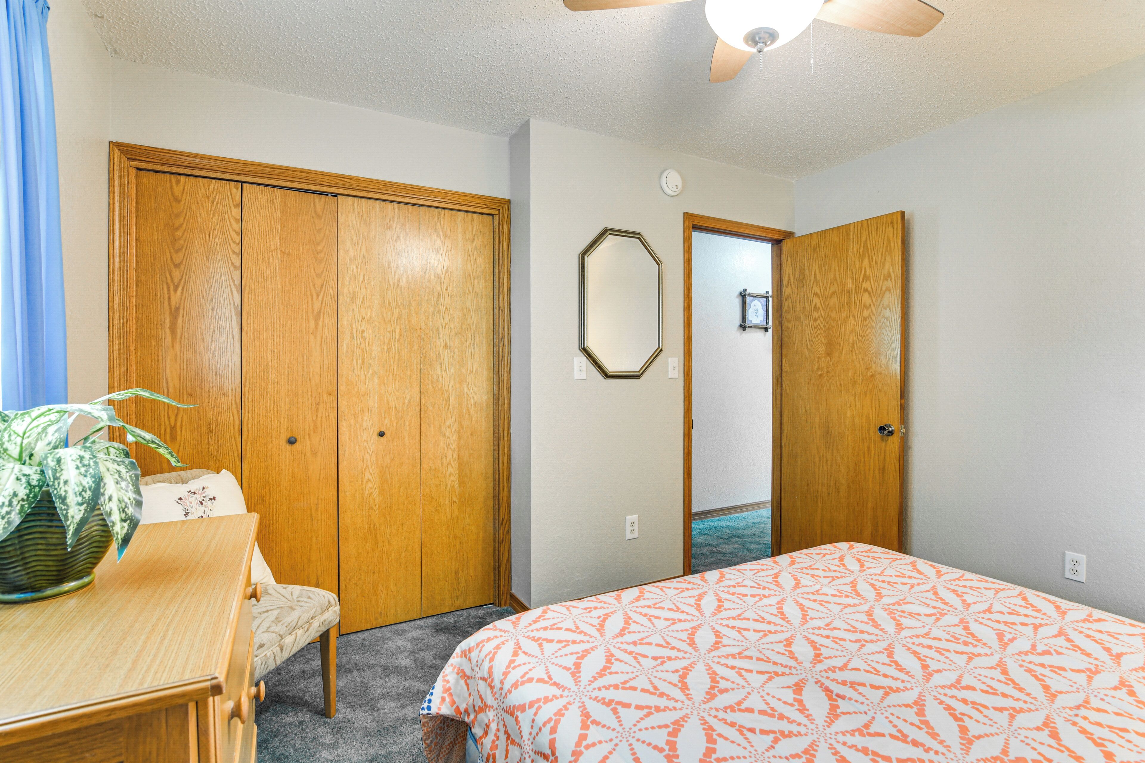 A bedroom with a bed, a wooden wardrobe, a desk with a chair, and a potted plant.