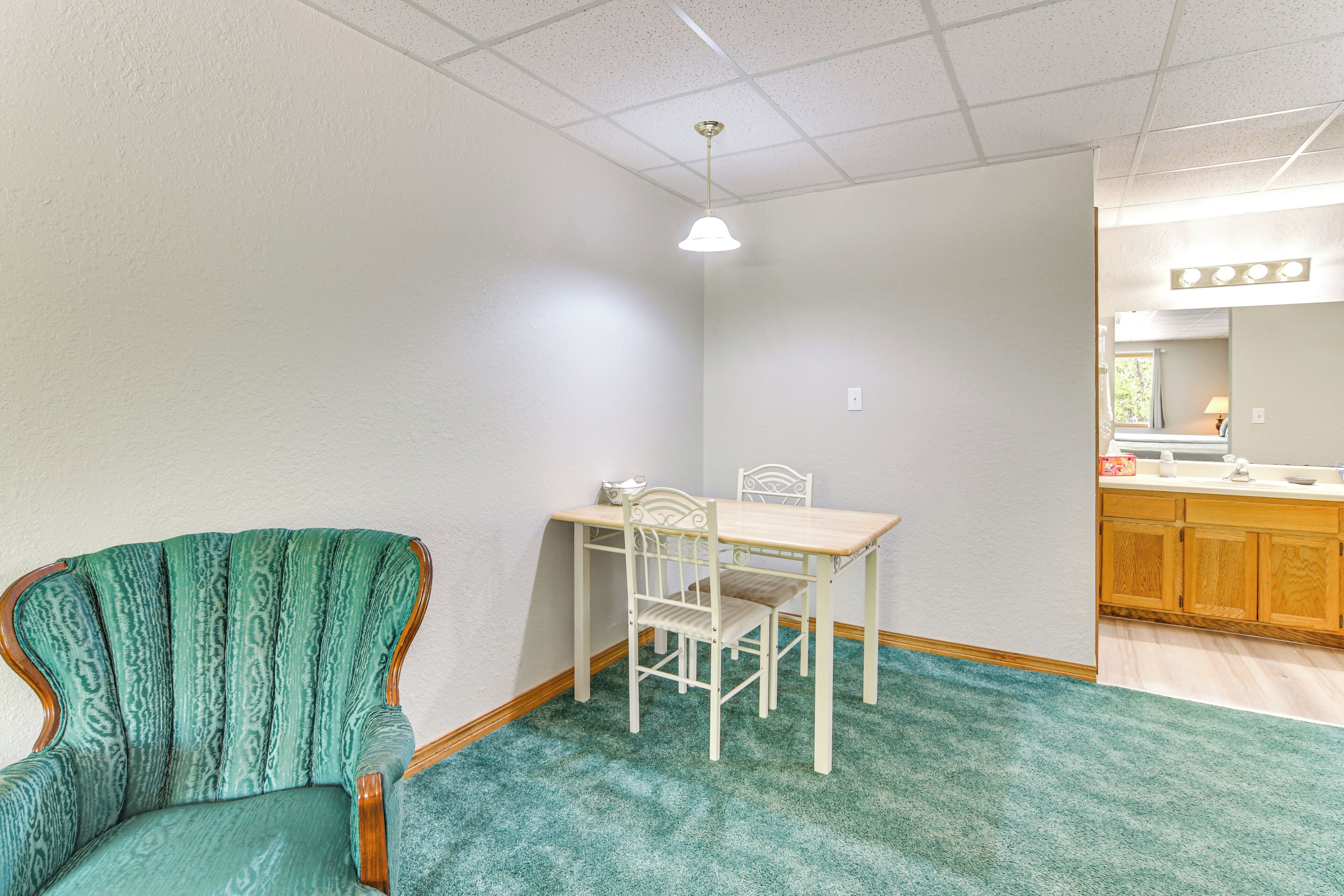 A room with a green armchair, a white dining table with two chairs, a kitchen area with wooden cabinets, and a ceiling with a grid pattern.
