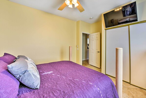 A bedroom with a purple bedspread, a ceiling fan, a TV mounted above the bed, and a wardrobe with a mirror.