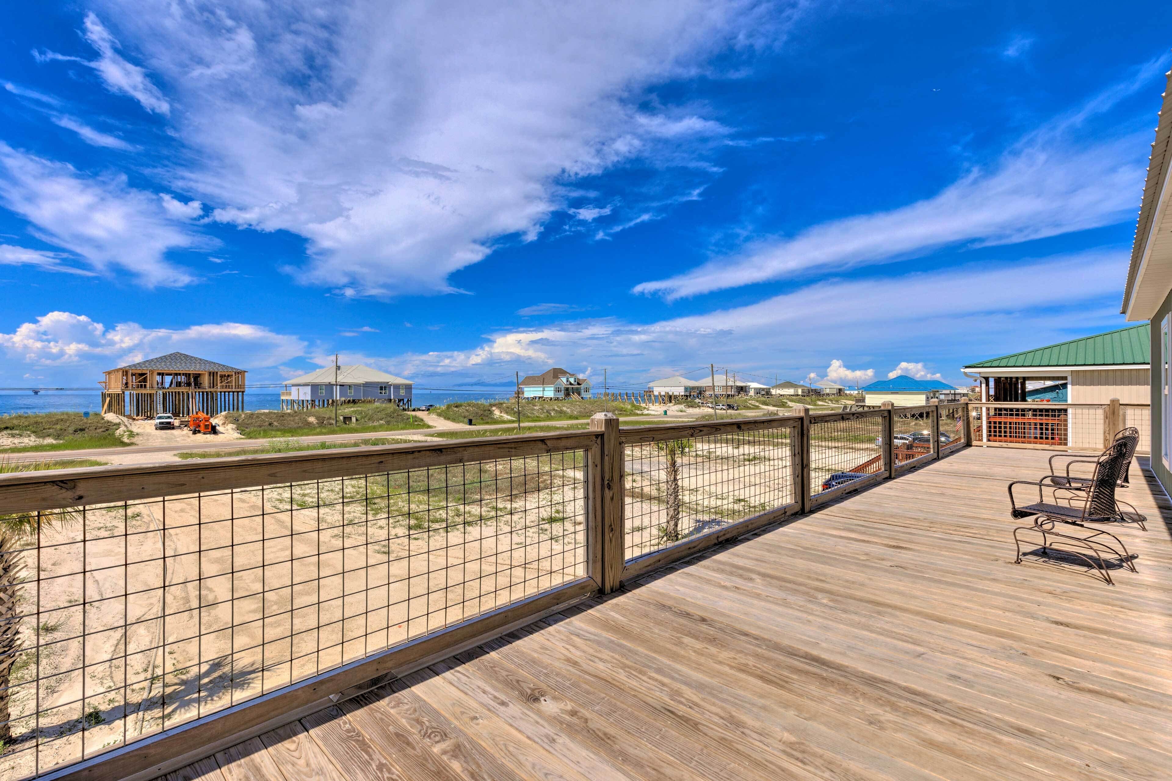 Una terraza de madera con barandilla metálica que da a una playa con edificios al fondo.
