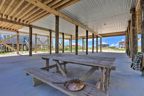 A covered picnic area with wooden benches and a table, under a metal roof, with a view of houses and beach in the background.