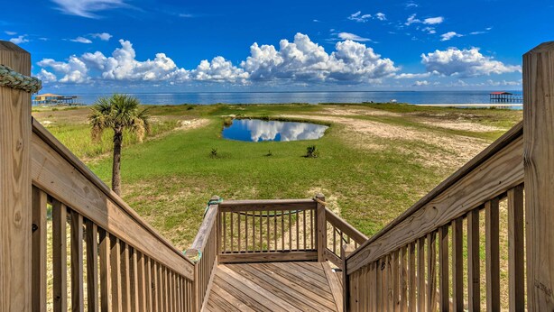 A wooden deck leading to a grassy area with a small pond, a palm tree, and a distant pier under a clear blue sky. - Bayfront Dauphin Island Home w/ Beach Access! (Dauphin Island)