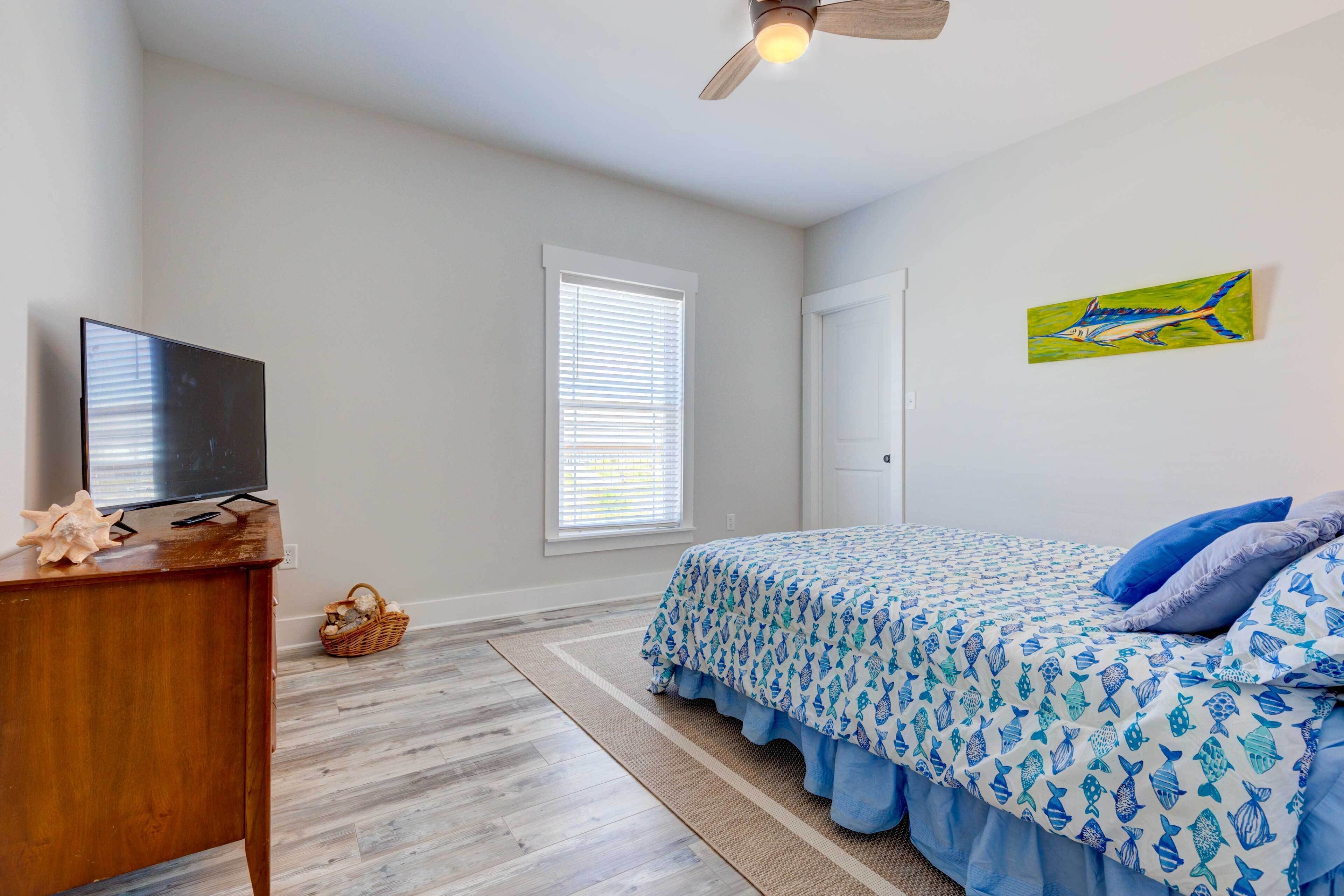 A bedroom with a bed, a television on a wooden dresser, a ceiling fan, a window with blinds, and a door.