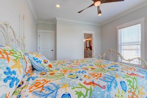 Une chambre à coucher avec un lit agrémenté d’une couvre-lit aux motifs colorés sur le thème de la mer, d’une tête de lit blanche et d’un ventilateur de plafond.