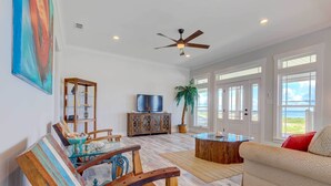 A spacious living room with a wooden coffee table, a sofa, and a television on a cabinet. There is a ceiling fan and a large window with a view. - Bayfront Dauphin Island Home w/ Beach Access! (Dauphin Island)