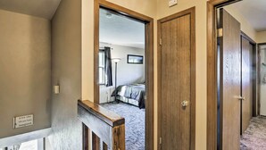 Un couloir avec des portes en bois et un sol recouvert de moquette, qui mène à une chambre dotée d’un lit et d’une fenêtre.