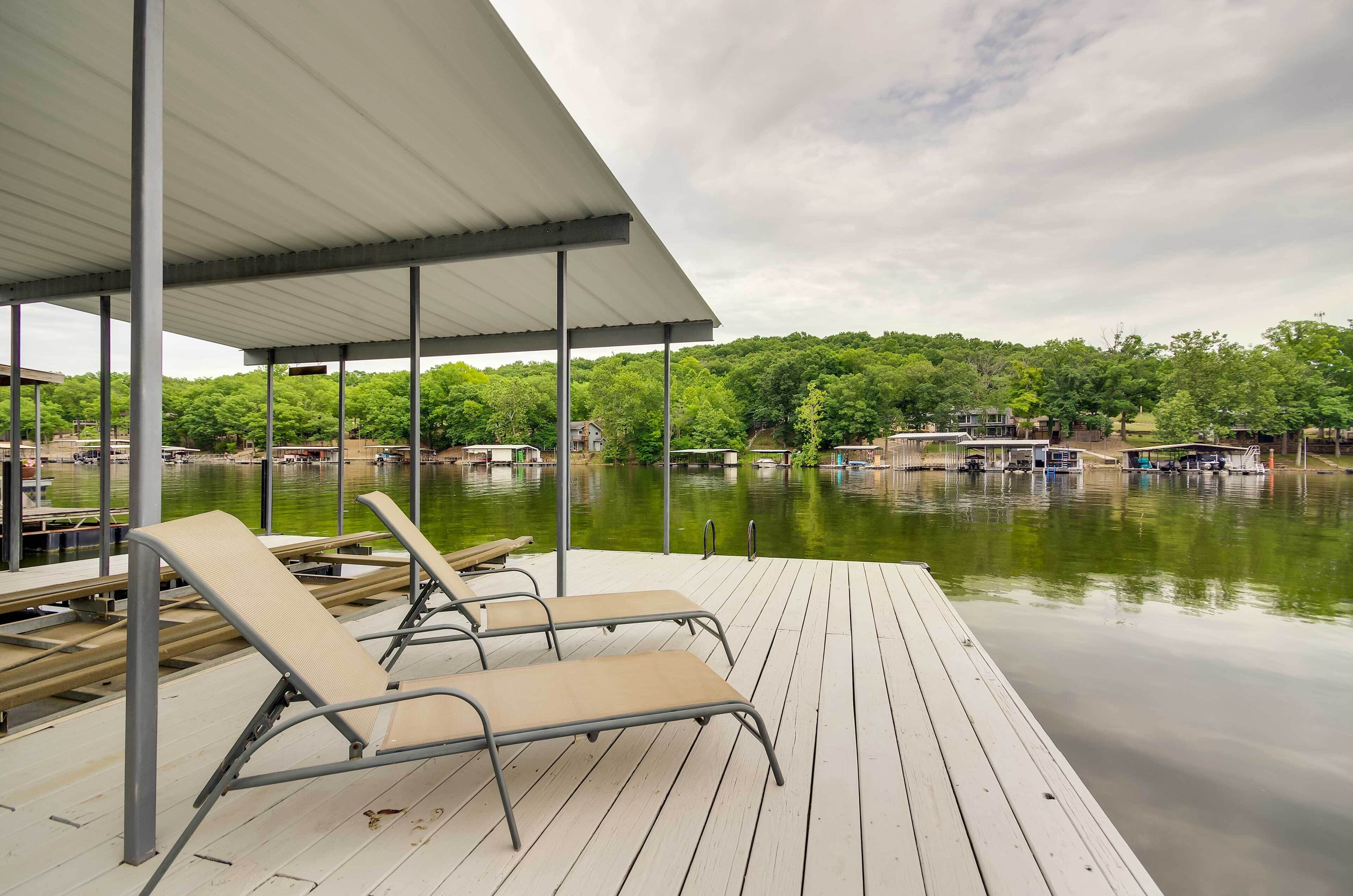 Una terraza de madera con sillones, un área techada y vistas a barcos en un lago tranquilo, con árboles al fondo.